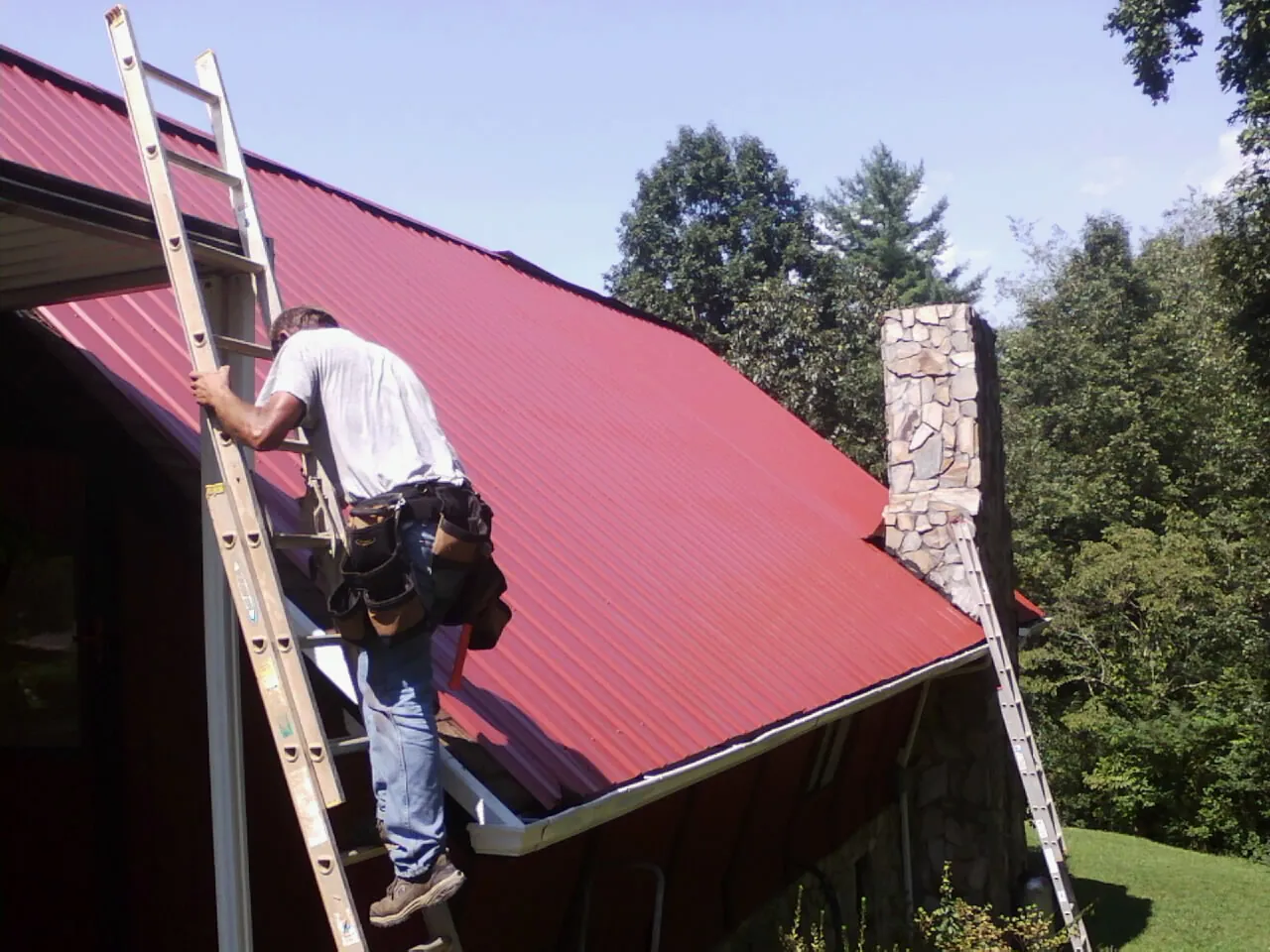 Roofer on a ladder beside a red metal roof and stone chimney — Soffit & Fascia Repair by Roofing Brothers in Orlinda
